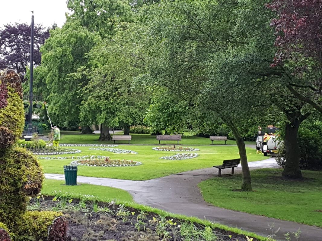 Lush green park with flower beds, benches, and a maintenance vehicle on a pathway under overcast skies.