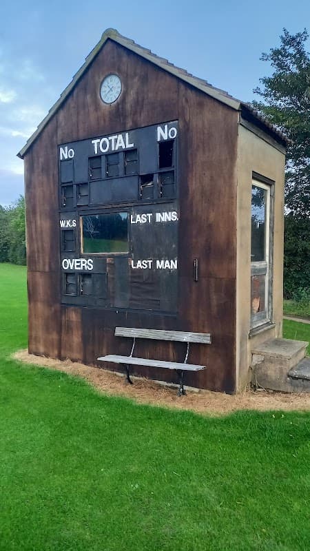 Weathered scoreboard building at Raskelf Cricket Club, with benches and a clock, set on a green cricket field.