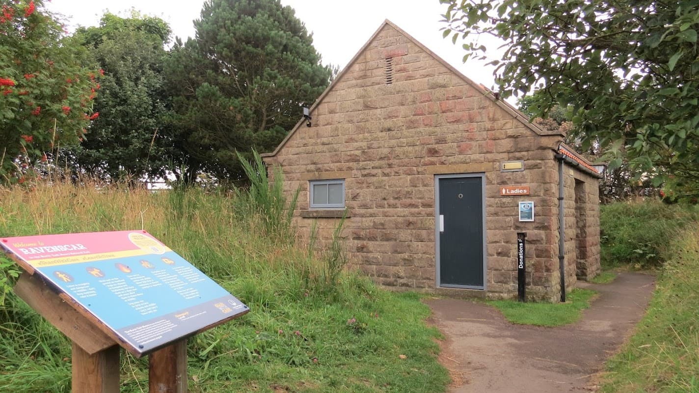 Stone building with a gray door, signage for ladies' toilets, and an information board in a grassy area.