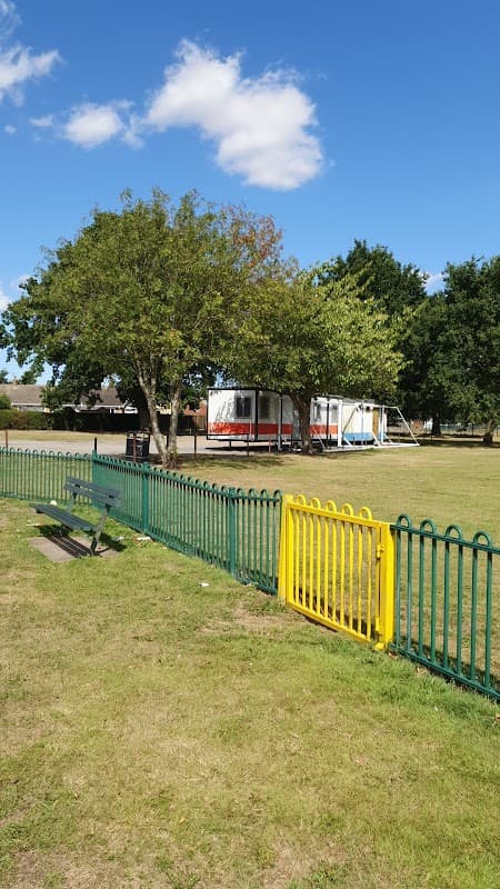 Football field with a green fence, a bench, trees, and a building in the background under a clear blue sky.