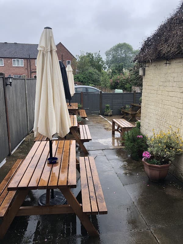Wooden picnic tables with umbrellas in a wet outdoor area, surrounded by greenery and a low fence.
