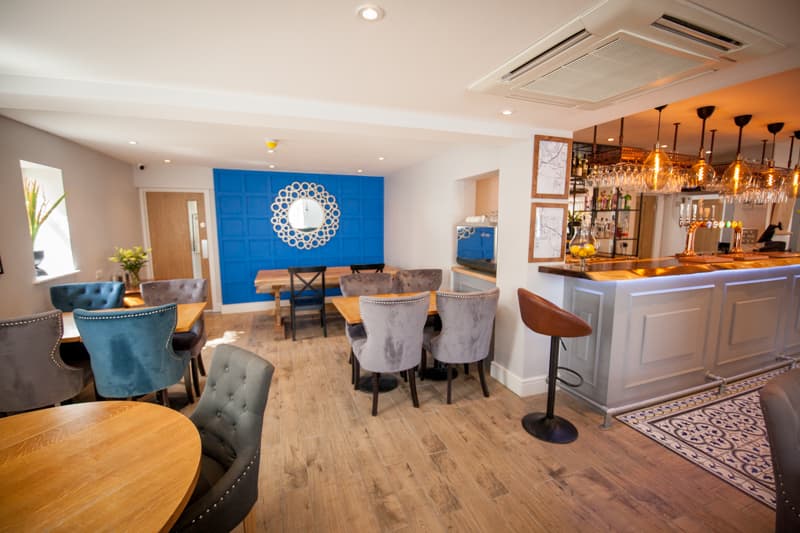 Stylish bar interior with blue accent wall, wooden tables, plush chairs, and a well-stocked bar area.