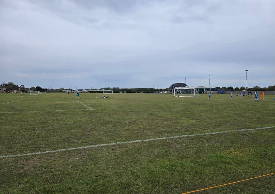 Expansive grassy field with soccer goals, players in blue jerseys, and a pavilion under a cloudy sky.