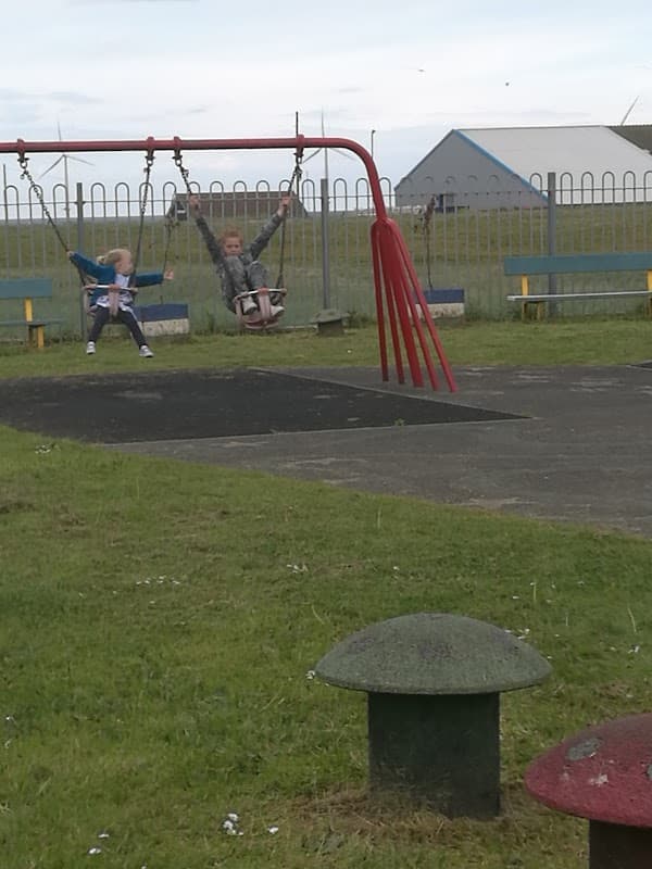 Two children swing on red swings in a grassy park with a blue sky and a building in the background.