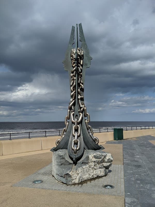Sculpture resembling chains and blades on a coastal promenade with a cloudy sky and sea in the background.