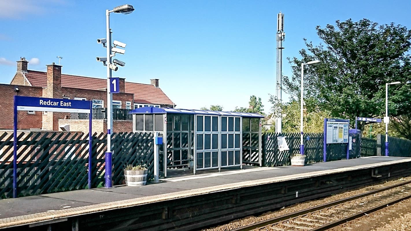 Redcar East station platform with a shelter, signage, and surrounding greenery under a clear blue sky.