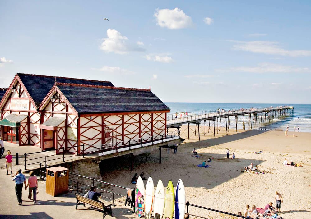Victorian-style building by the beach, with a pier extending into the sea and surfers' boards lined up in the foreground.