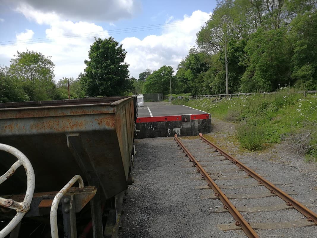 Rusty train car on tracks with a platform in the background, surrounded by greenery and trees under a cloudy sky.