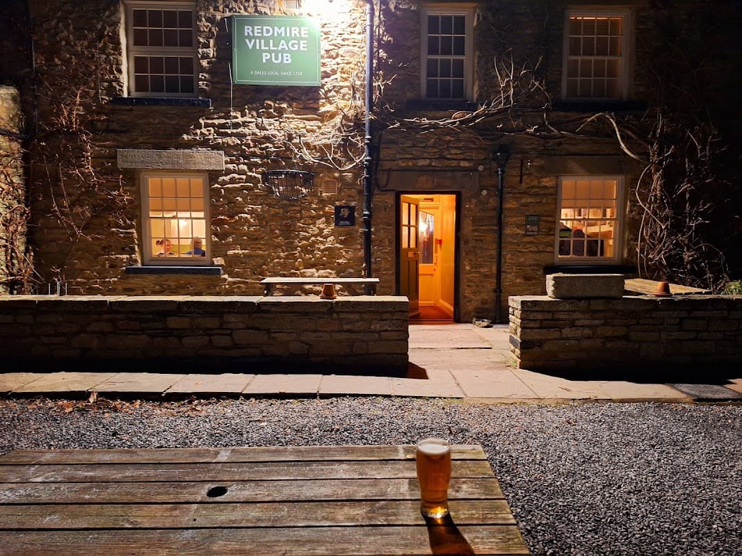 Redmire Village Pub with stone exterior, warm lighting, and a pint of beer on a wooden table in the foreground.