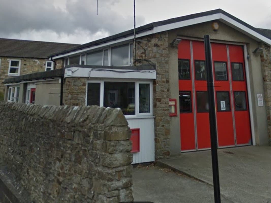 Reeth Fire Station features stone walls, large red garage doors, and a white entrance, set against a cloudy sky.