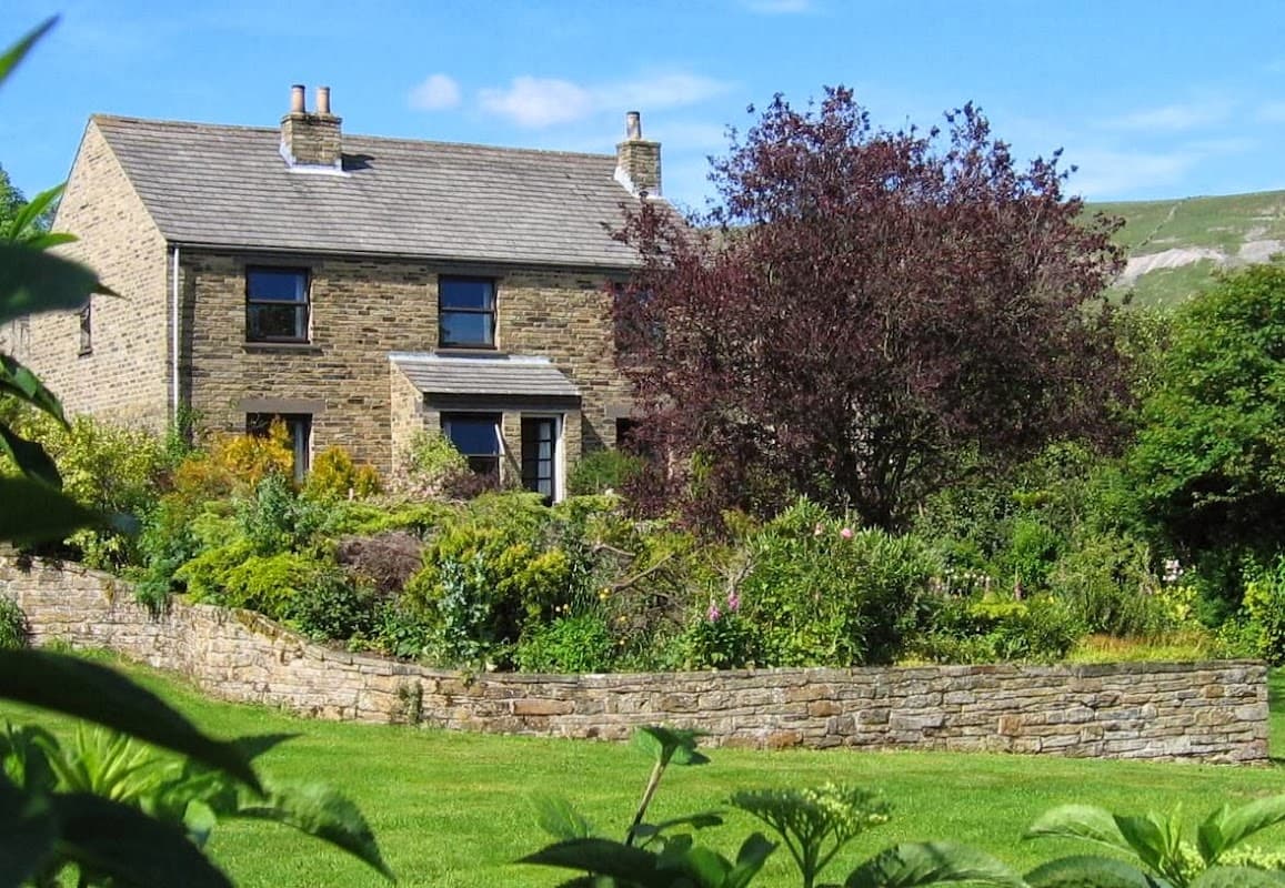 Stone cottage surrounded by lush gardens and a low stone wall, with a vibrant tree and rolling hills in the background.