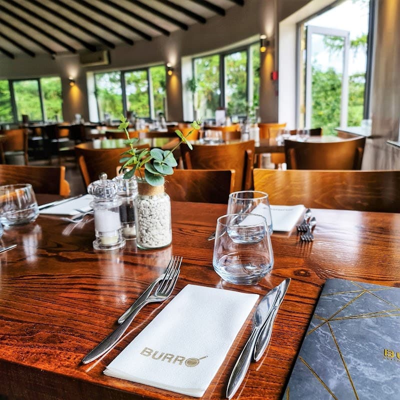 Elegant dining setup at Burro Restaurant, featuring wooden tables, glassware, and a view of greenery through large windows.