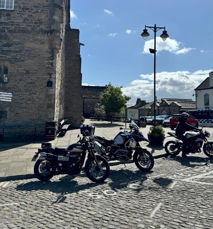 Motorcycles parked in a cobbled area with a pay and display sign, surrounded by buildings and a clear blue sky.
