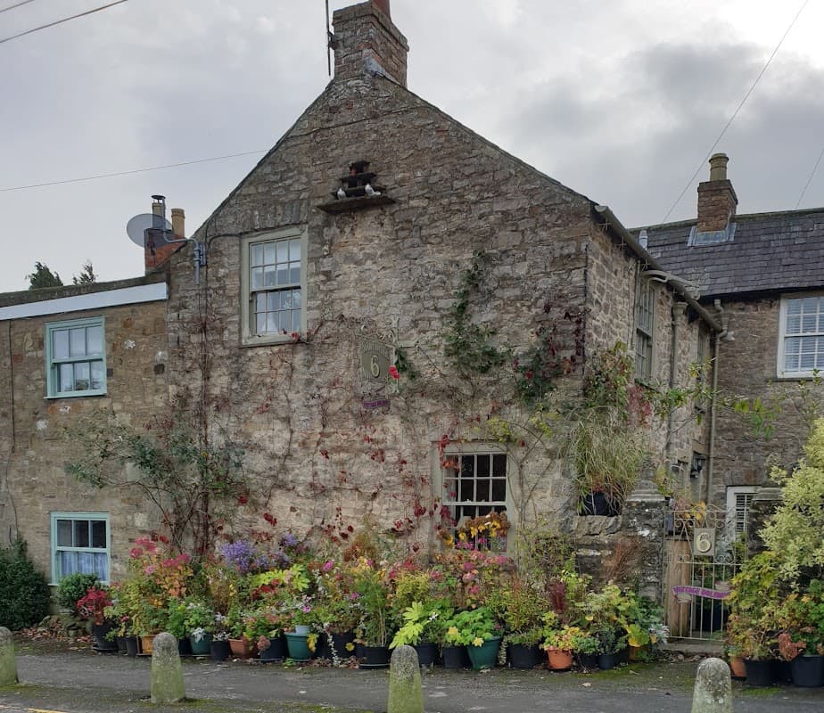 Stone cottage with colorful flower pots, ivy, and a house number 6, set against a cloudy sky.
