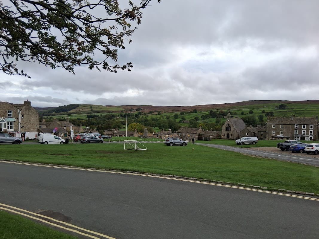 Pay & Display parking area with cars, grassy space, stone buildings, and a backdrop of hills under a cloudy sky.