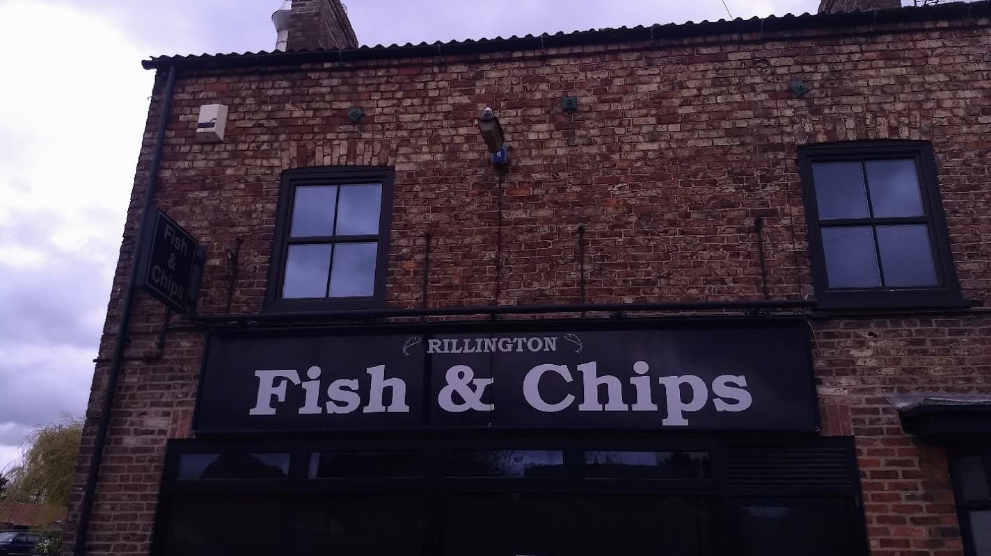 Brick building with a sign reading "Rillington Fish & Chips" and windows above. Cloudy sky in the background.