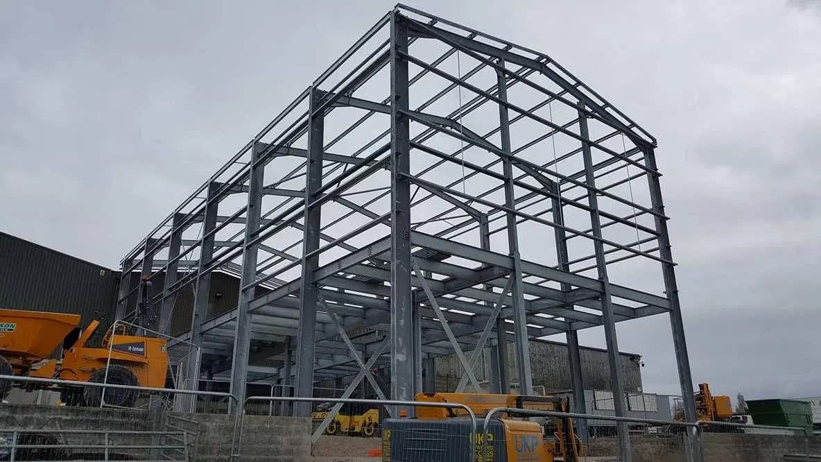 Steel framework of a building under construction, with machinery and equipment nearby. Overcast sky in the background.