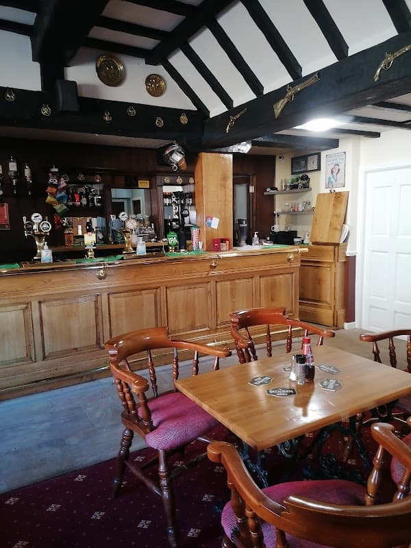 Wooden bar with taps, stools, and tables; rustic decor featuring dark beams and framed pictures in a cozy pub setting.