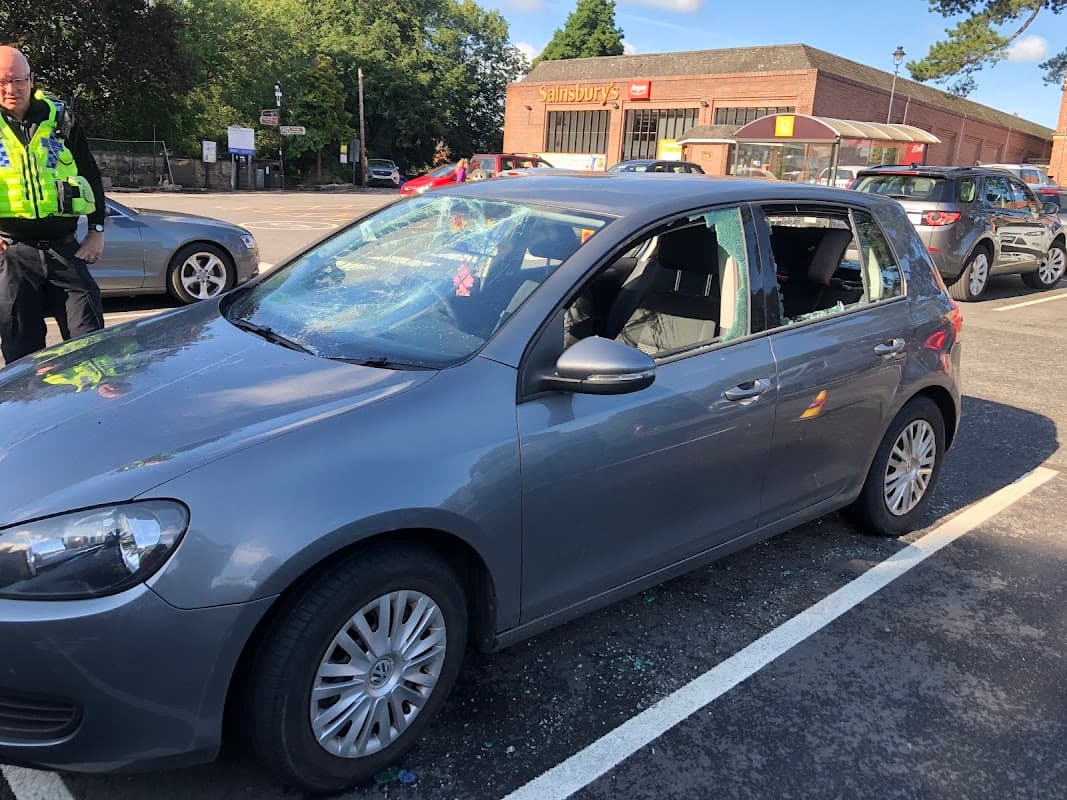A gray car with shattered windows parked in a lot, with a police officer nearby and shops in the background.