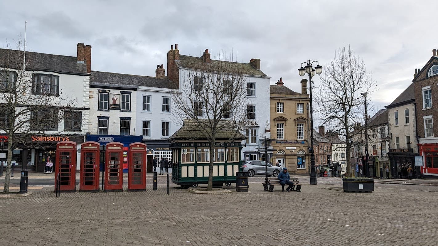 Historic market square with red phone boxes, a green kiosk, and surrounding shops in Ripon, Yorkshire.