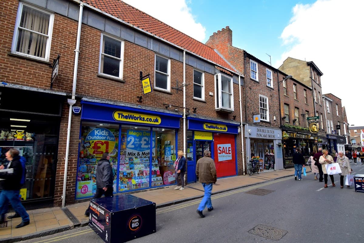 Colorful storefront of The Works bookshop, featuring books and toys, with pedestrians walking on a cobblestone street.