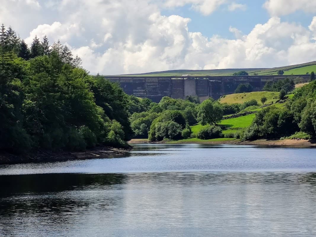 Ryburn Reservoir with lush greenery, calm water, and a dam in the background under a partly cloudy sky.