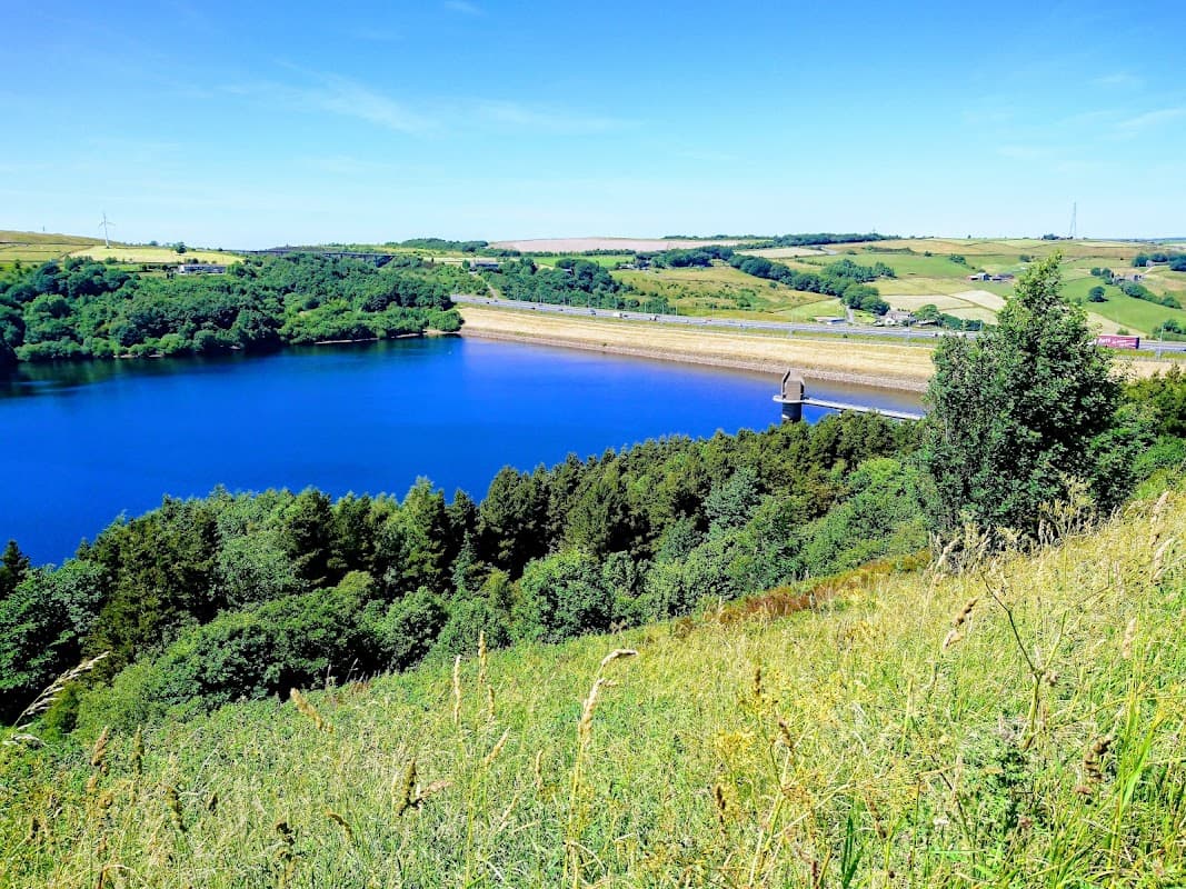 Scammonden Water with a blue sky, green hills, and a dam in the distance, surrounded by lush vegetation.