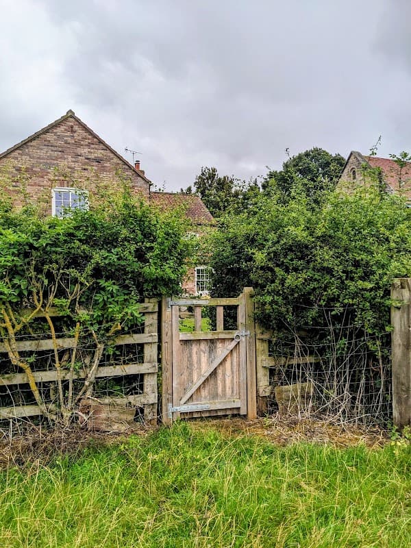 A wooden gate set in a hedge, leading to a stone building partially obscured by greenery and cloudy skies.