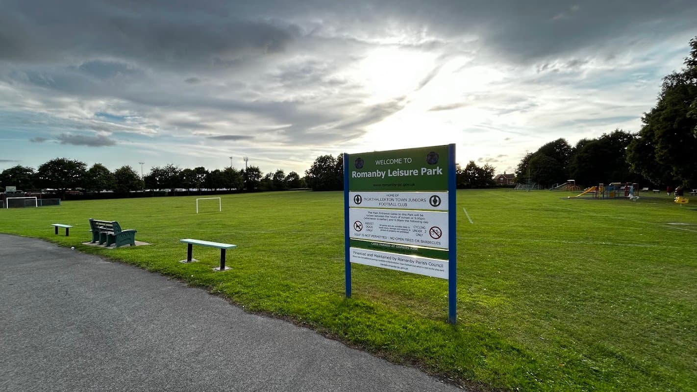 Welcome sign for Romanby Leisure Park with green grass, benches, and cloudy sky in the background.