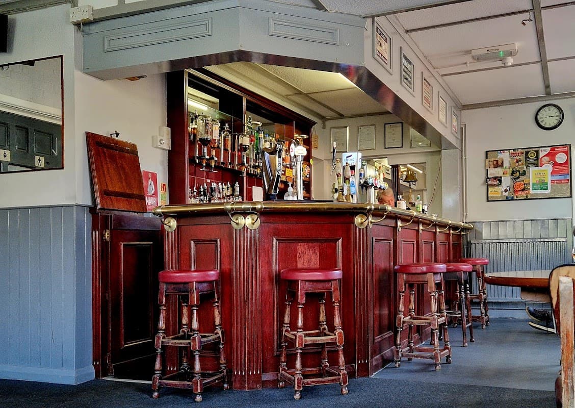 Wooden bar with red stools, mirrors, and various drink taps; walls adorned with notices and a clock.