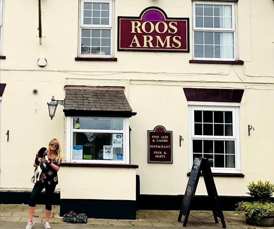 Historic hotel facade with "ROOS ARMS" sign, a woman holding a dog, and a blackboard menu outside.