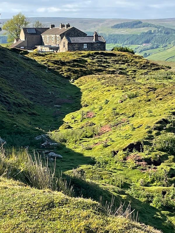 Historic stone building on a hillside, surrounded by lush greenery and rolling hills under a clear blue sky.