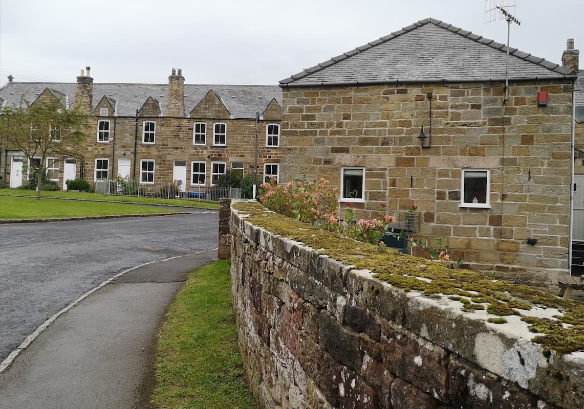 Stone building with a sloped roof, surrounded by greenery and a moss-covered wall, in a quaint village setting.