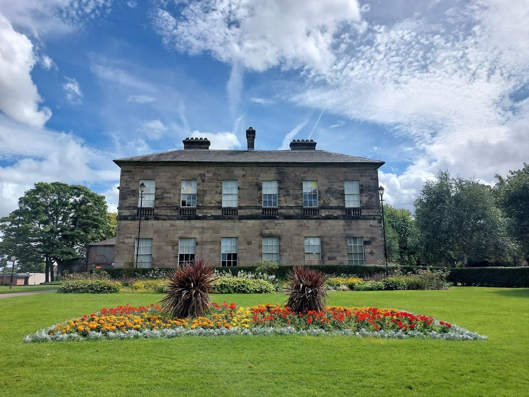 Historic stone building with large windows, surrounded by vibrant flower beds and lush green lawns under a blue sky.