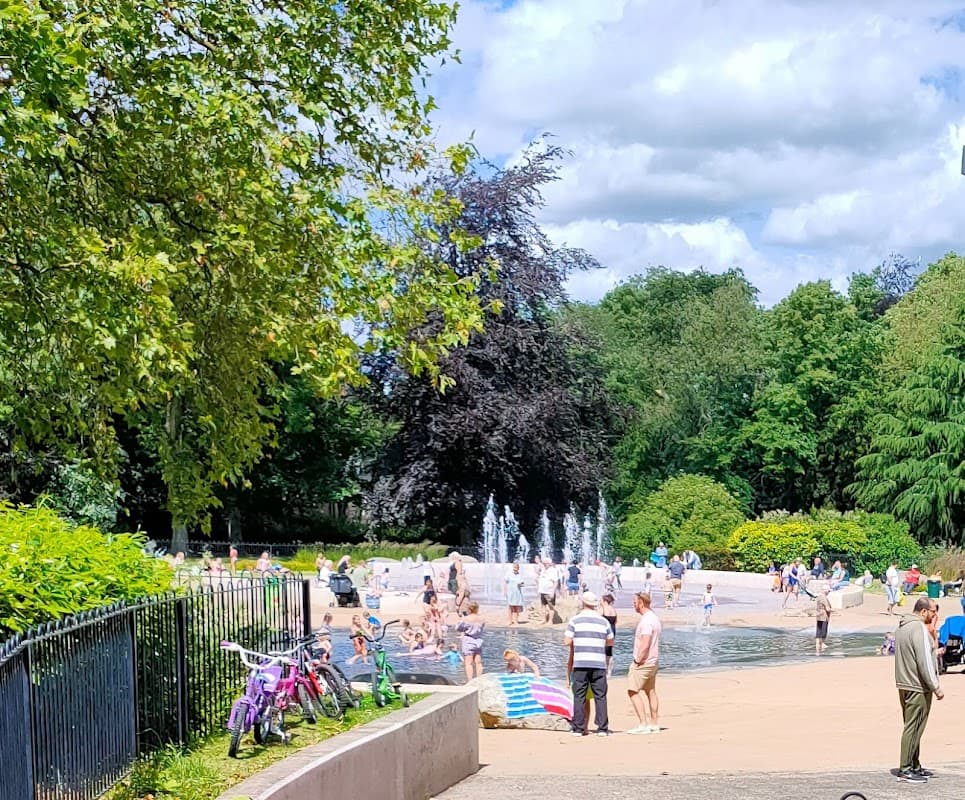 Families enjoying a sunny day at Clifton Park, with a splash pad, greenery, and bicycles nearby.