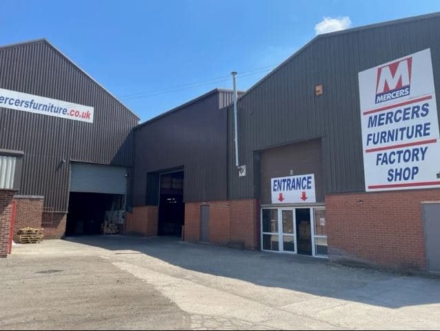 Mercers Furniture Ltd factory shop entrance with large signage, brick facade, and industrial buildings under a blue sky.
