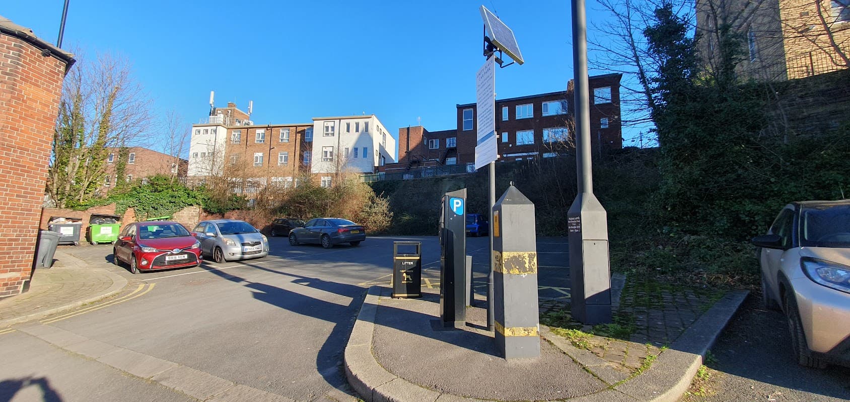 Pay & Display car park with parked cars, a payment machine, and buildings in the background under a clear blue sky.