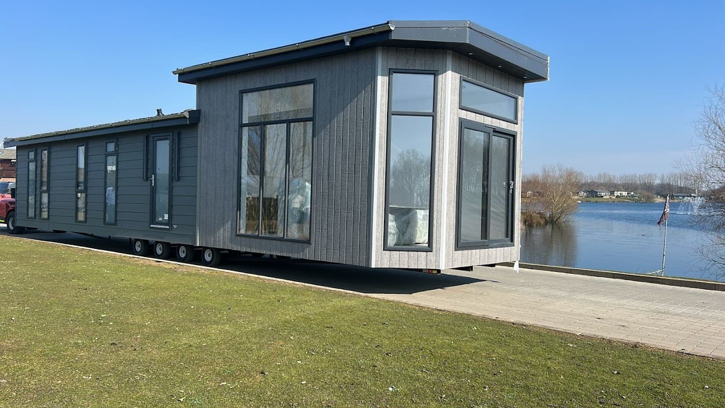 Modern gray mobile home with large windows, situated by a serene lake in Routh, Yorkshire, under a clear blue sky.