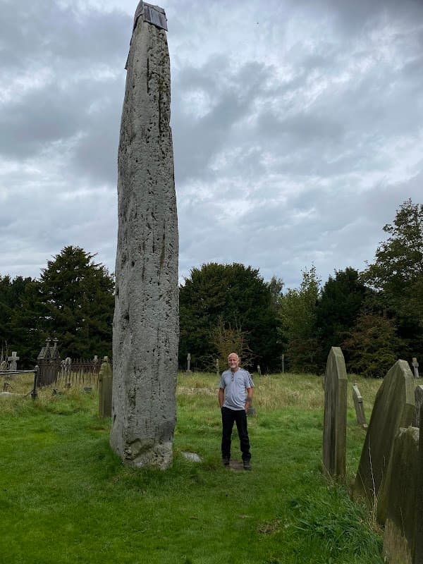 Tall, weathered stone monolith surrounded by grass and gravestones, with a person standing nearby under a cloudy sky.