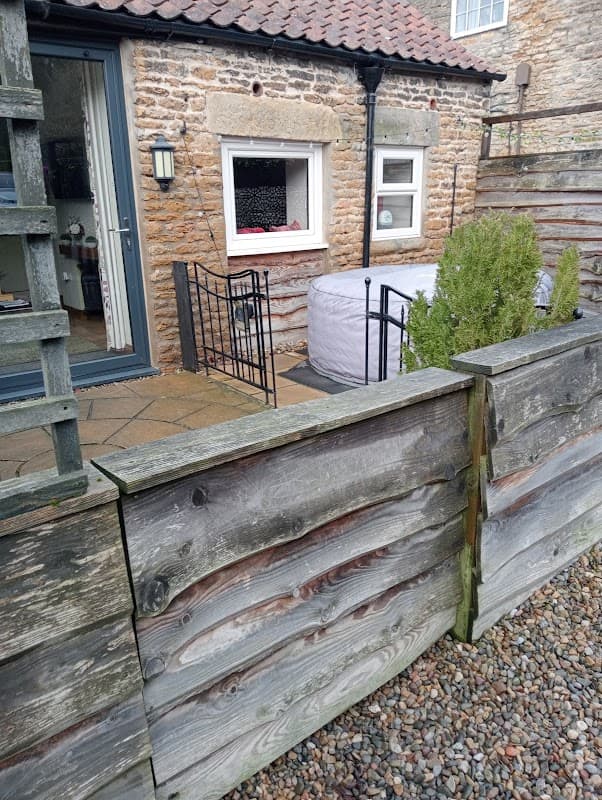 Rustic stone cottage with a slate roof, surrounded by a wooden fence and a small garden area.