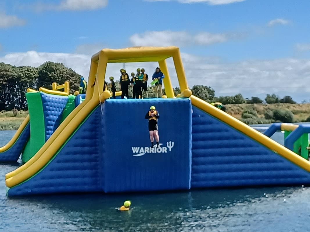 Colorful inflatable water obstacle course with people in helmets, jumping and swimming in a lake under a clear blue sky.