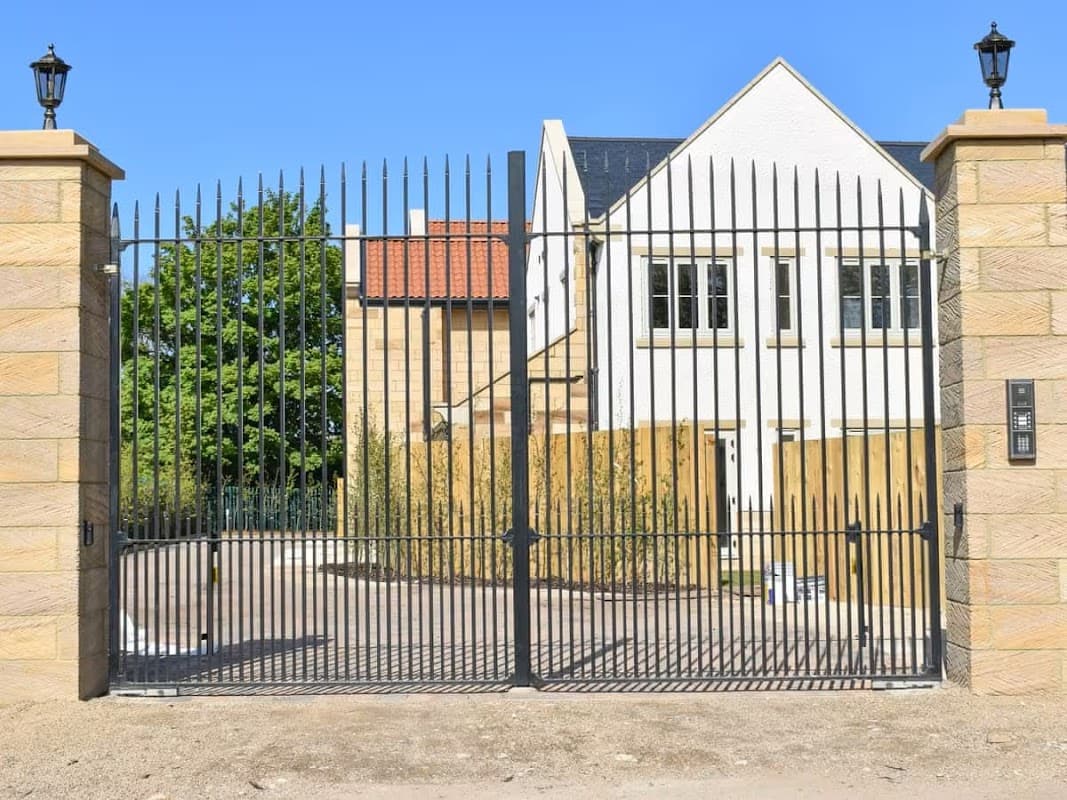 Black wrought iron gates with stone pillars, leading to a modern building with a white facade and blue roof.