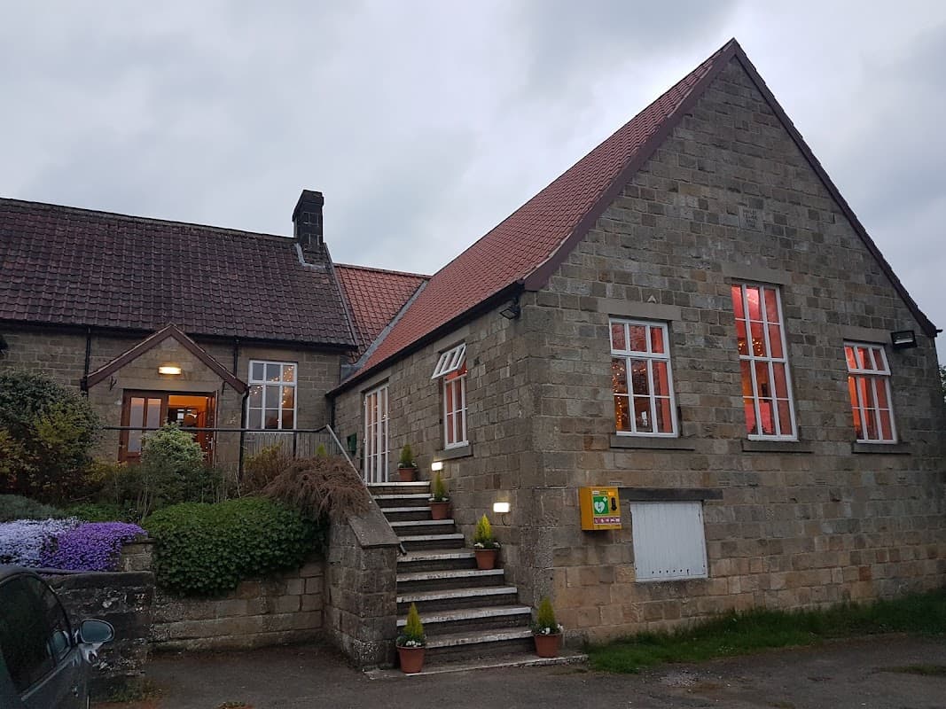Stone building with red roof, steps leading to entrance, and colorful flowers in front, under a cloudy sky.