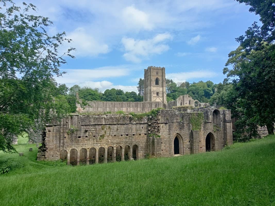 Ruins of Fountains Abbey surrounded by lush greenery under a blue sky with scattered clouds.