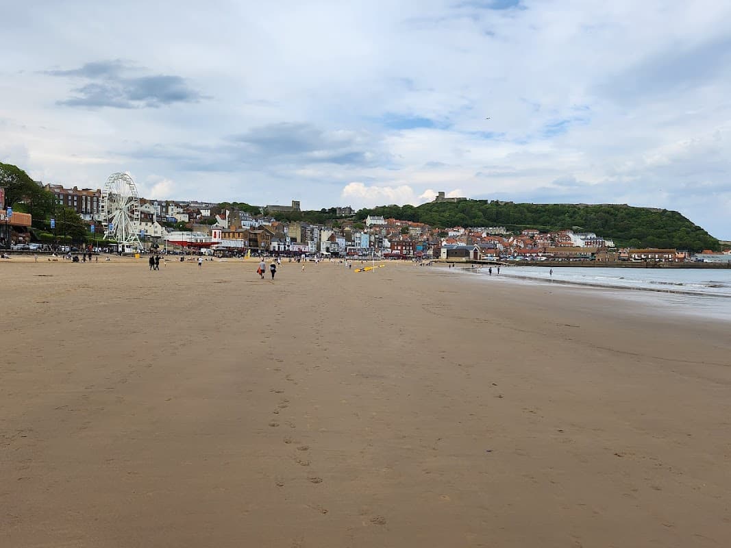 Beach scene with sandy shore, distant amusement rides, and Scarborough's coastal buildings under a cloudy sky.