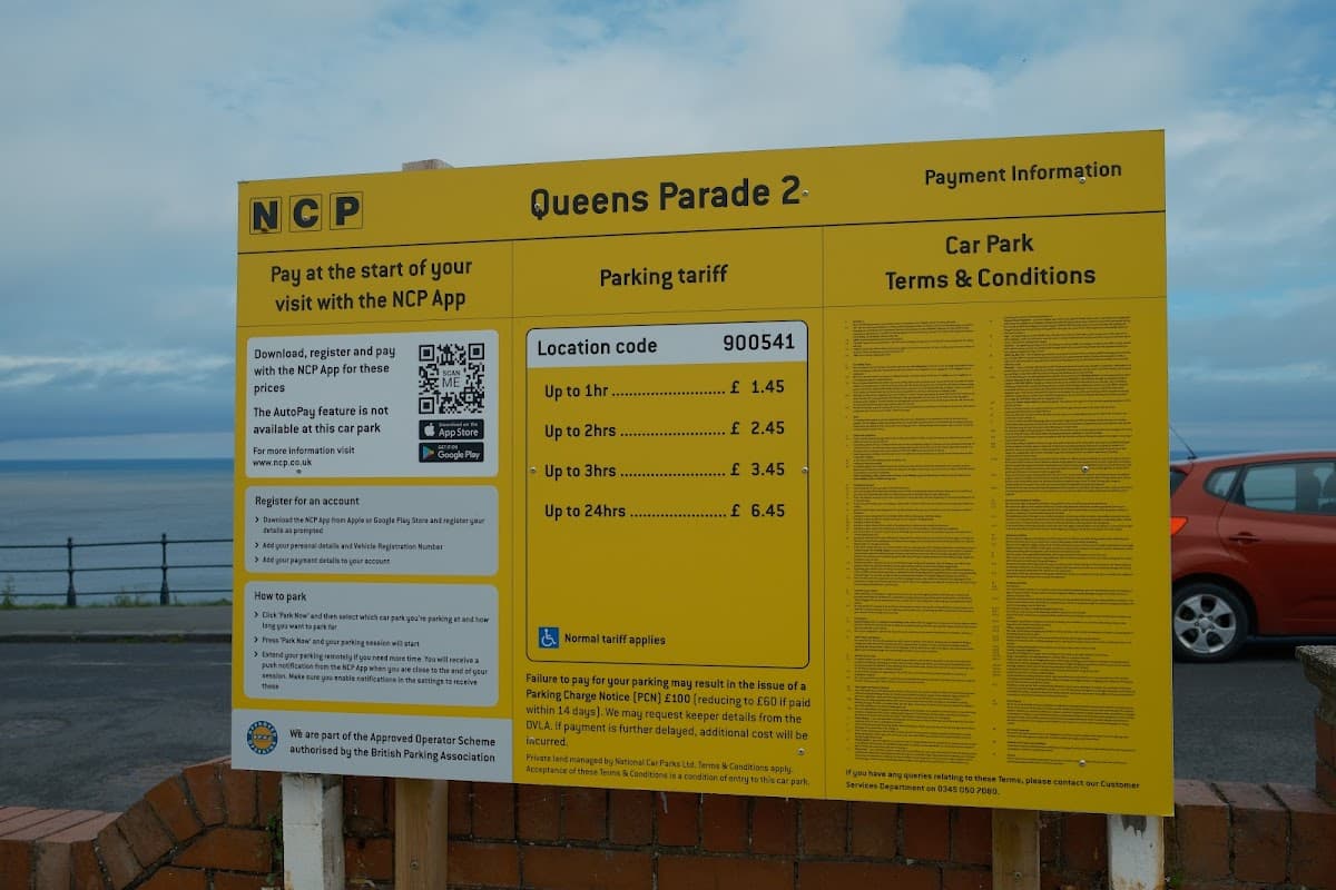 Bright yellow sign displaying parking information and tariffs for NCP Queens Parade 2, with a coastal view in the background.