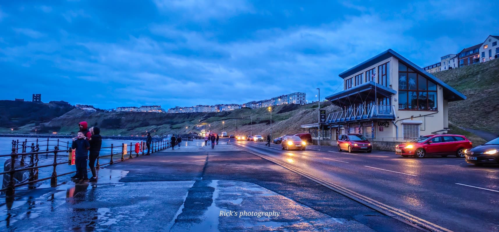 Pay & Display parking near the sea, with cars, pedestrians, and a building under a cloudy blue sky in Scarborough.