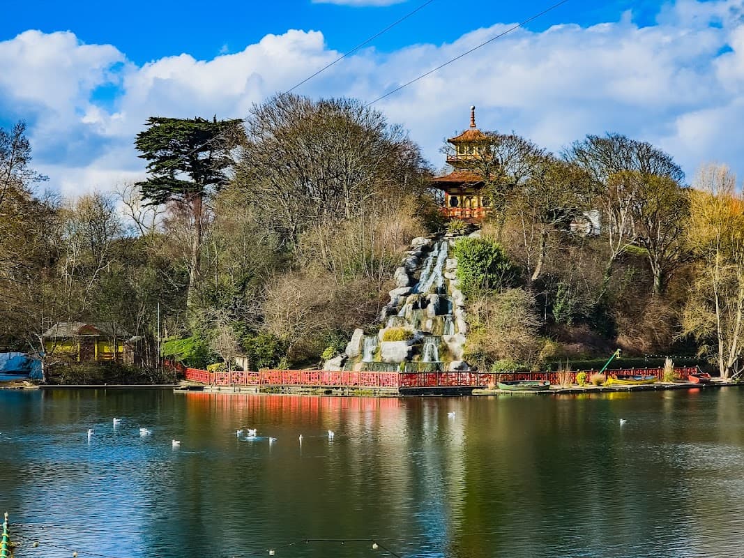 Serene lake with a waterfall, surrounded by trees and a pagoda, under a blue sky with fluffy clouds.