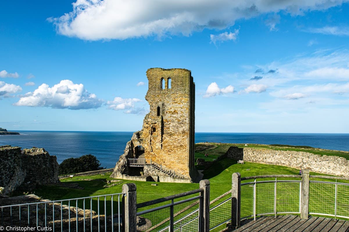 Ruins of Scarborough Castle stand against a blue sky, overlooking the sea and green landscape.