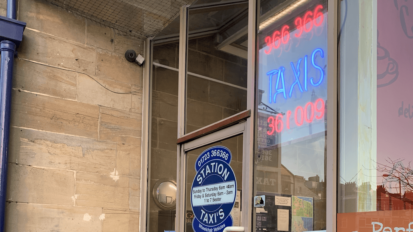Bright neon "TAXIS" sign and a blue circular sign for "Station Taxis" at a hire center entrance in Scarborough.